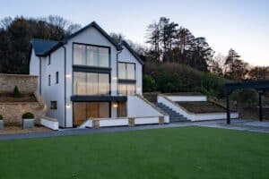 A modern two-story house with slimline sliding doors and large glass windows, surrounded by greenery. It features an outdoor staircase and a neatly maintained lawn.