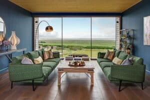A modern living room with slimline sliding doors, two green sofas, a wooden coffee table, and a large floor-to-ceiling window overlooking a grassy landscape. The walls are painted dark blue.