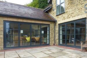 Stone house exterior featuring large art deco steel frame doors that lead to an open-plan interior with a dining table and colorful chairs. The traditional patio area is paved with stones.
