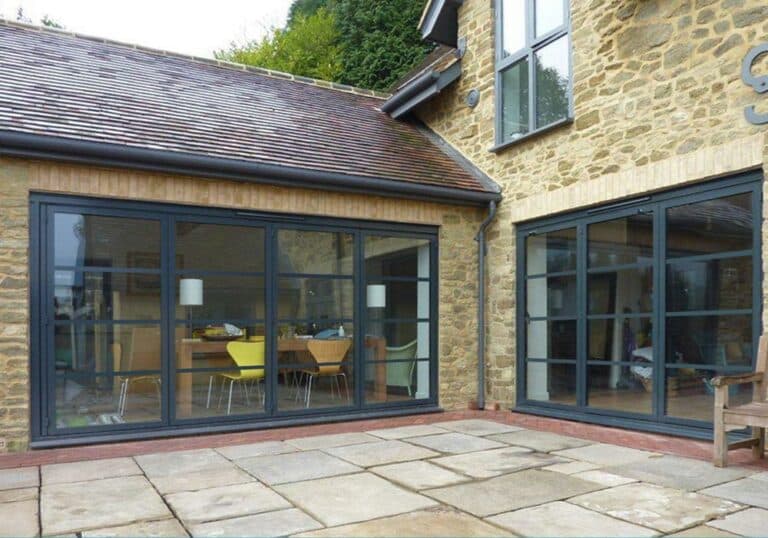 Stone house exterior featuring large art deco steel frame doors that lead to an open-plan interior with a dining table and colorful chairs. The traditional patio area is paved with stones.