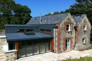 A stone and brick house with a modern metal and glass extension, featuring steel frame windows, is set against a backdrop of trees under a blue sky.
