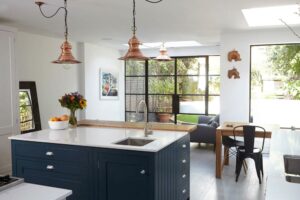 Modern kitchen with a navy island, copper pendant lights, and a sleek dining area. Large steel frame windows allow natural light to flood in. Decor includes a fruit bowl, flowers, and wall art that add an Art Deco touch.