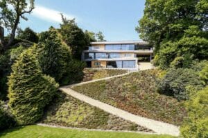 Modern hillside house with large windows and sleek aluminium sliding doors, surrounded by lush trees and a sloped garden. A stone path zigzags through the landscaped greenery.