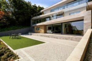 Modern three-story house with large windows and sleek aluminum sliding doors, surrounded by trees. Features a spacious patio with outdoor seating and a neatly trimmed lawn.