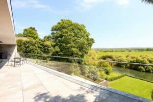 Spacious terrace with aluminium sliding doors and glass railing overlooks a green landscape and a river on a sunny day.