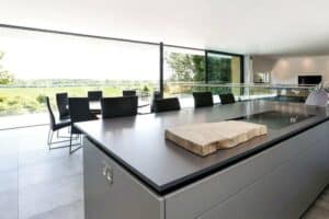 Modern kitchen with sleek gray countertops, large island, and aluminium sliding doors leading to glass walls overlooking a scenic outdoor view. Dining area with dark chairs in the background.