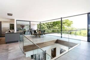 Modern open-plan kitchen and dining area with aluminium sliding doors that create seamless glass walls overlooking the landscape. Features a central island, dining table, and stunning views of greenery.