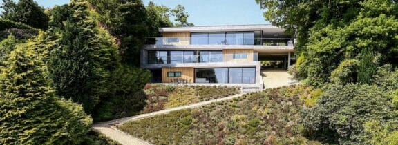 Modern multi-story house with large glass windows and sleek aluminium sliding doors, surrounded by trees and greenery. A sloped garden leads to the entrance.