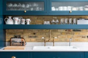 A kitchen with blue cabinets, exposed brick backsplash, and a white countertop is complemented by brass faucets. Aluminium heritage doors add charm, while a toaster sits beside shelves holding cups and glasses.