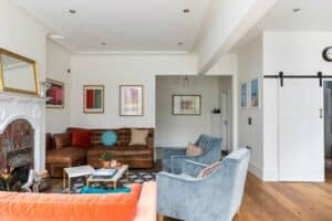 A cozy living room features a brown leather sofa, blue armchairs, and a white fireplace framed by colourful art. The room, with aluminium heritage doors enhancing its charm, boasts wooden floors and a mix of modern and classic decor.