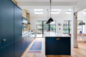 Modern kitchen with blue cabinets, a long island, and pendant lights. Wooden flooring and a colorful rug lead to a dining area featuring large windows, skylights, and elegant aluminium heritage doors.