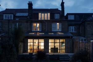 A three-story brick house with illuminated windows at dusk, featuring a modern glass extension on the ground floor adorned with elegant aluminium heritage doors.