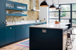 A modern kitchen with blue cabinets, a brick backsplash, and a central island with stools. Pendant lights hang above, while large windows framed by aluminium heritage doors offer a view of the garden.