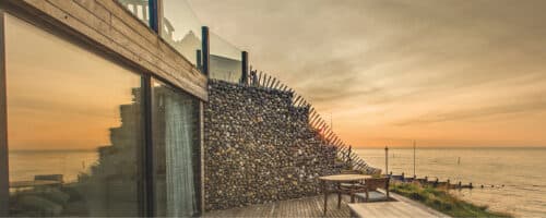 Modern building with a stone facade and glass railing, featuring sleek glass pocket sliding doors that open to overlook the ocean at sunset. A small outdoor table and chairs are on the deck, inviting relaxation as the day fades away.