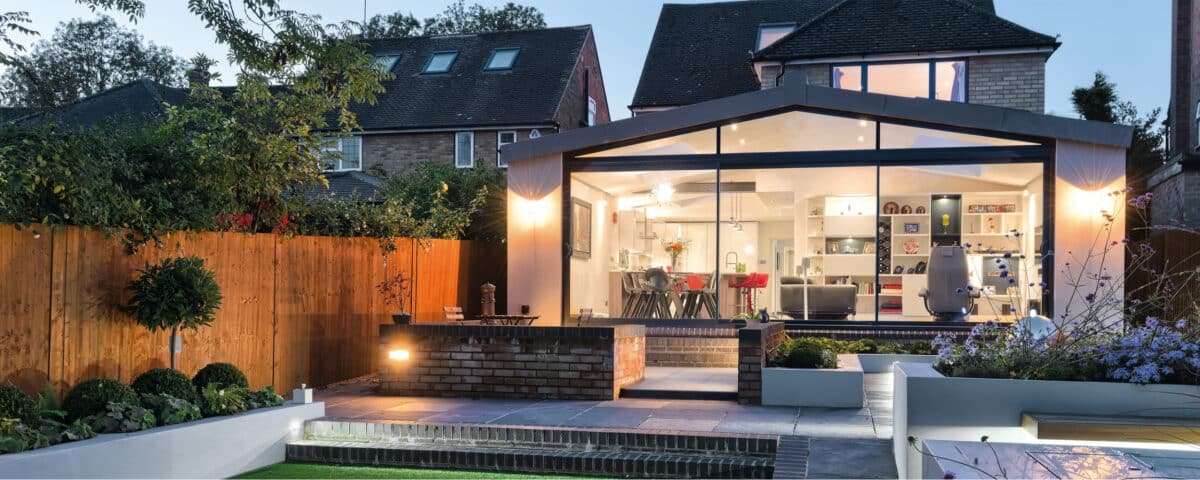 Modern home with large glass windows and a well-lit, open interior. The exterior features a tiled patio, garden, and wooden fence. Aluminium sliding doors seamlessly connect indoor and outdoor spaces, set in a suburban neighborhood at dusk.