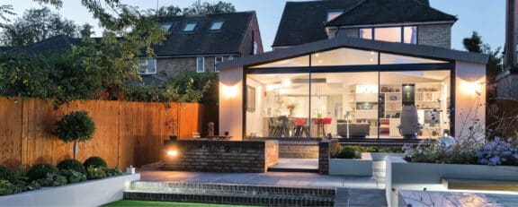 Modern home with large glass windows and a well-lit, open interior. The exterior features a tiled patio, garden, and wooden fence. Aluminium sliding doors seamlessly connect indoor and outdoor spaces, set in a suburban neighborhood at dusk.