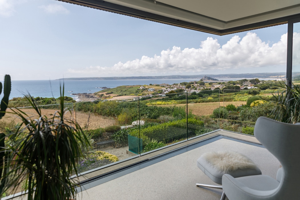 Modern room with large glass windows and corner sliding doors, offering a panoramic view of fields, coastline, and blue sky with clouds; lounge chair and plants in the foreground.