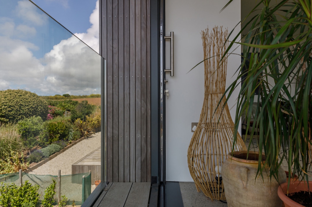 A modern entryway featuring corner sliding doors with a large glass panel, wooden siding, potted plants, and a decorative wicker piece, overlooking a landscaped garden under a partly cloudy sky.