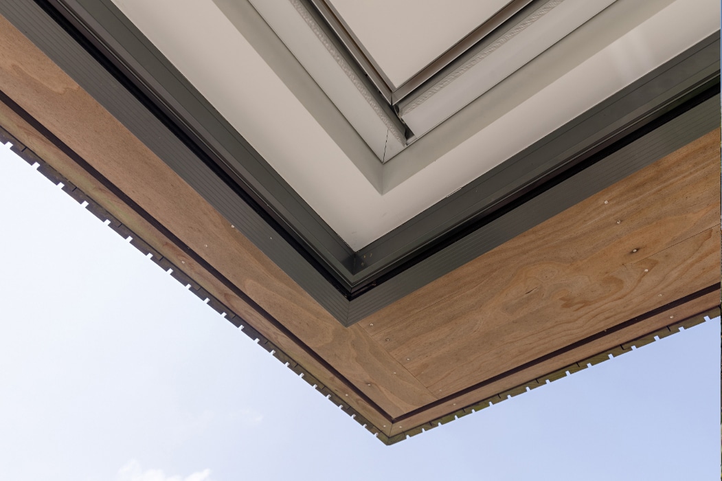 Close-up view of the underside of a modern building’s corner roof with plywood paneling, metal framing, and sleek corner sliding doors, set against a clear blue sky.