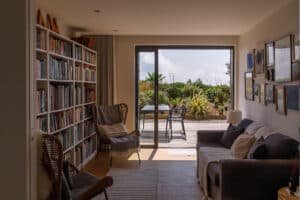 A cozy living room with bookshelves, chairs, and a sofa features corner sliding doors that open onto a sunny patio with a table and chairs, surrounded by lush plants and greenery.