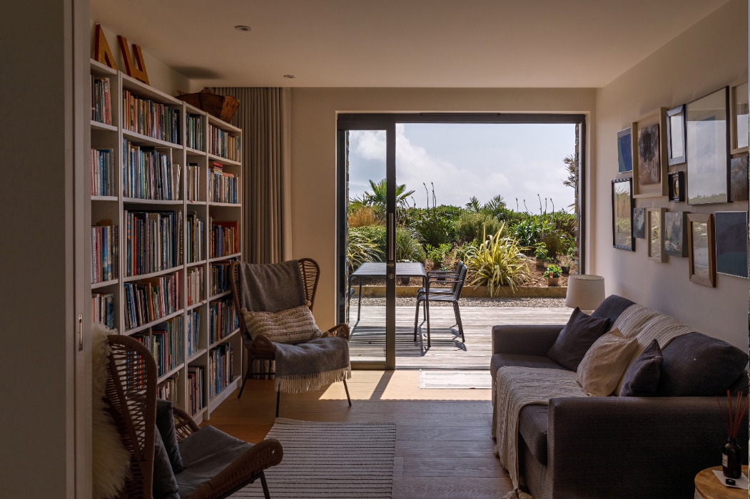 A cozy living room with bookshelves, chairs, and a sofa features corner sliding doors that open onto a sunny patio with a table and chairs, surrounded by lush plants and greenery.