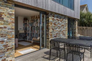 Modern house with stone exterior, corner sliding doors, visible bookshelf inside, and a black outdoor dining table and chairs on a wooden deck.