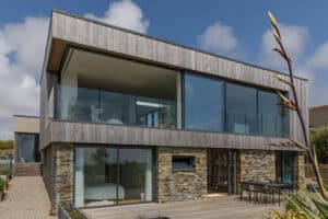 A modern two-story house with large glass windows, corner sliding doors, a stone and wood exterior, an upper balcony, and an outdoor dining area under a partly cloudy sky.