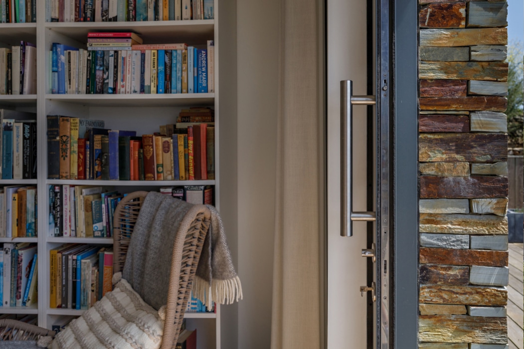 A woven chair with a striped cushion and blanket is placed next to a bookshelf filled with books, near an open door with a metal handle, stone wall, and elegant corner sliding doors.