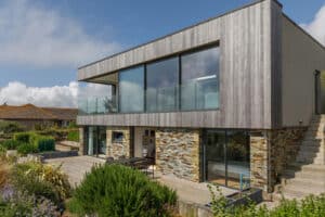 Modern two-story house with large glass windows, stone and wood exterior, balcony, outdoor patio, and sleek corner sliding doors, surrounded by landscaped plants under a partly cloudy sky.