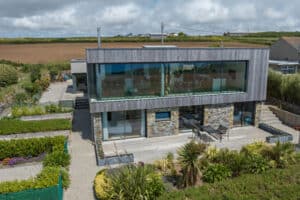 Modern two-story house with large glass windows, stone accents, and landscaped garden, featuring corner sliding doors, situated in a rural area with open fields in the background.