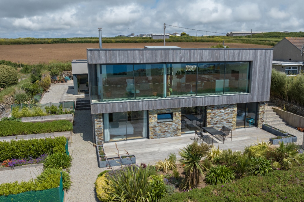 Modern two-story house with large glass windows, stone accents, and landscaped garden, featuring corner sliding doors, situated in a rural area with open fields in the background.