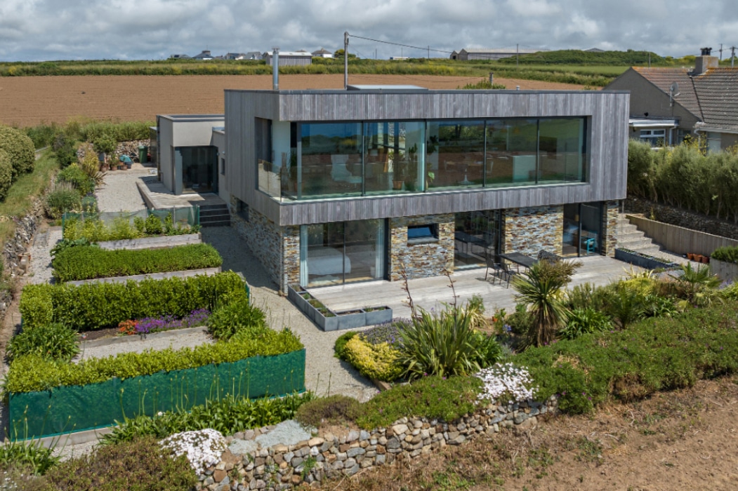 Modern two-story house with large glass windows and corner sliding doors, stone and wood exterior, surrounded by landscaped gardens and fields under a partly cloudy sky.