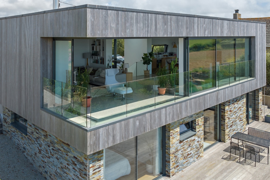 A modern two-story house with wood paneling, large glass windows, corner sliding doors, a glass balcony, and a stone base, overlooking an outdoor patio with seating.