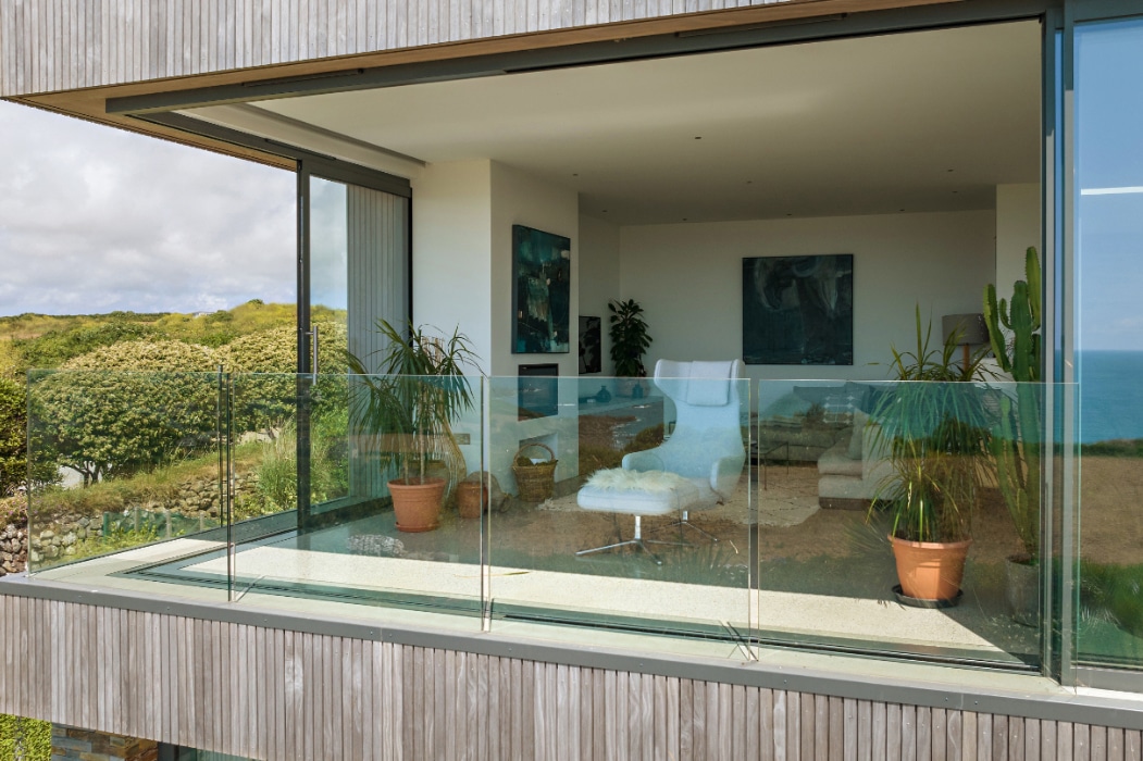 Modern living room with large glass windows and corner sliding doors, a balcony featuring glass railing, potted plants, and a view of greenery and the sea in the background.