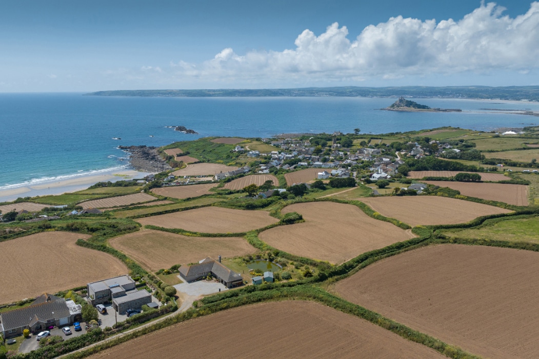 Aerial view of a coastal village with fields, scattered houses featuring corner sliding doors, and an island with a distinct structure in the distance, all framed by the sea and blue sky.