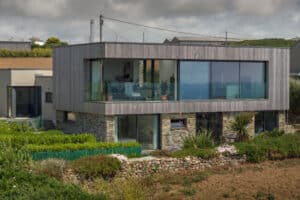 Modern two-story house with large glass windows and corner sliding doors, featuring a wooden upper facade and stone lower level, surrounded by greenery and a stone wall in a rural setting.