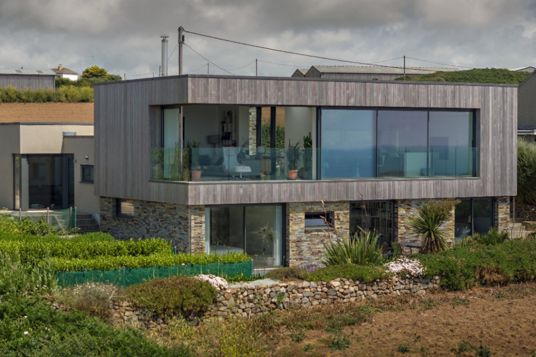 Modern two-story house with large glass windows and corner sliding doors, featuring a wooden upper facade and stone lower level, surrounded by greenery and a stone wall in a rural setting.