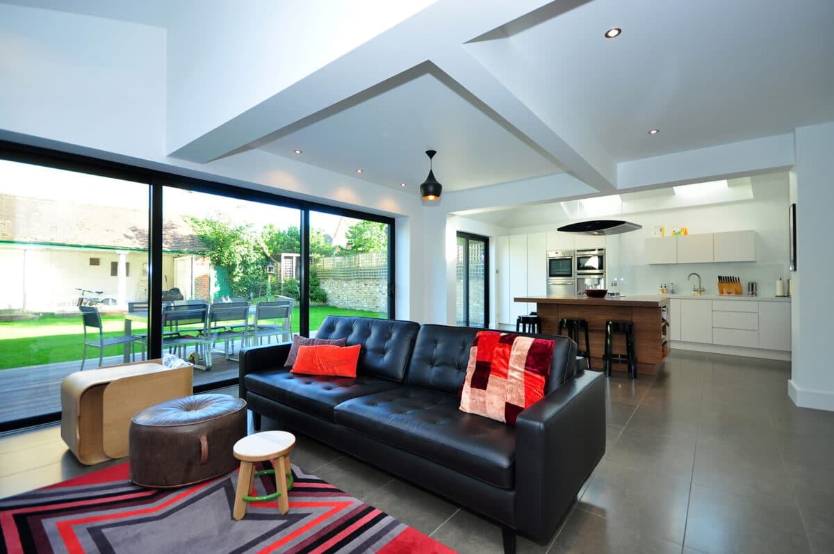 Modern open-plan living space with large sliding patio doors opening to garden, featuring black leather sofa and contemporary kitchen.