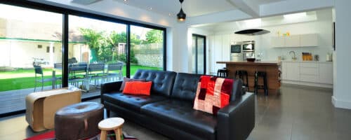 Modern open-plan living space with external glazed doors opening onto a garden patio area, featuring slim-profile aluminium frames and maximised natural light.