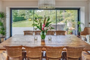 Dining room with a large wooden table, wicker chairs, and a vase of flowers. Glass doors lead to a grassy outdoor garden area with patio furniture.