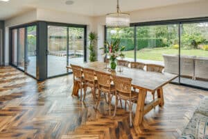 Spacious dining area with a wooden table, wicker chairs, herringbone wood floor, and large glass doors overlooking a garden with a patio.