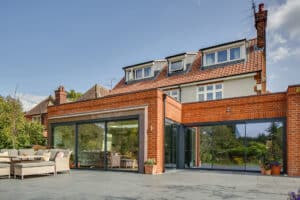 A two-story brick house boasts large aluminium bifold doors leading to the patio seating, offering a stunning garden view under a clear blue sky.