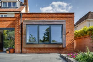 Modern brick house with large windows reflecting the sky and surrounding greenery, adjacent to a paved patio and a small garden with flowers.
