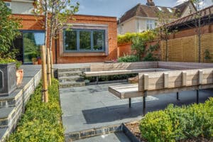 Modern backyard with a wooden bench, slate patio, and raised brick plant beds, surrounded by a privacy fence and greenery.