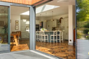 Modern kitchen and dining area with wooden herringbone floor, large island, pendant lights, and wicker chairs, viewed through open glass doors, with a garden visible outside.
