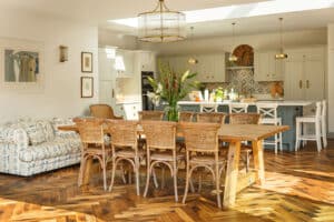 Spacious dining area with a wooden table and wicker chairs, a floral centerpiece, herringbone wood flooring, a sitting area with a patterned couch, and a kitchen in the background.