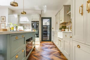 A modern kitchen with light gray cabinetry, brass handles, herringbone wood flooring, and island. Natural light from a window highlights the white countertop and decorative backsplash.