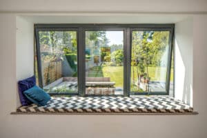 A window seat with a chevron-patterned cushion and two pillows overlooks a garden with trees and a lawn.