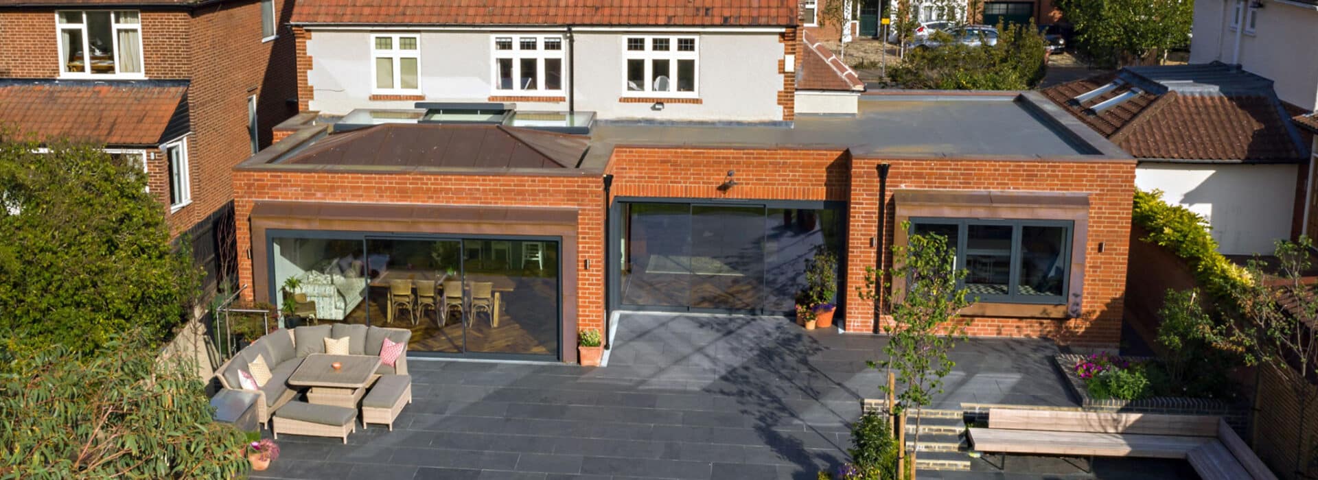 A modern brick house with sleek sliding aluminium doors opens onto a spacious tiled patio featuring outdoor seating and scattered greenery.