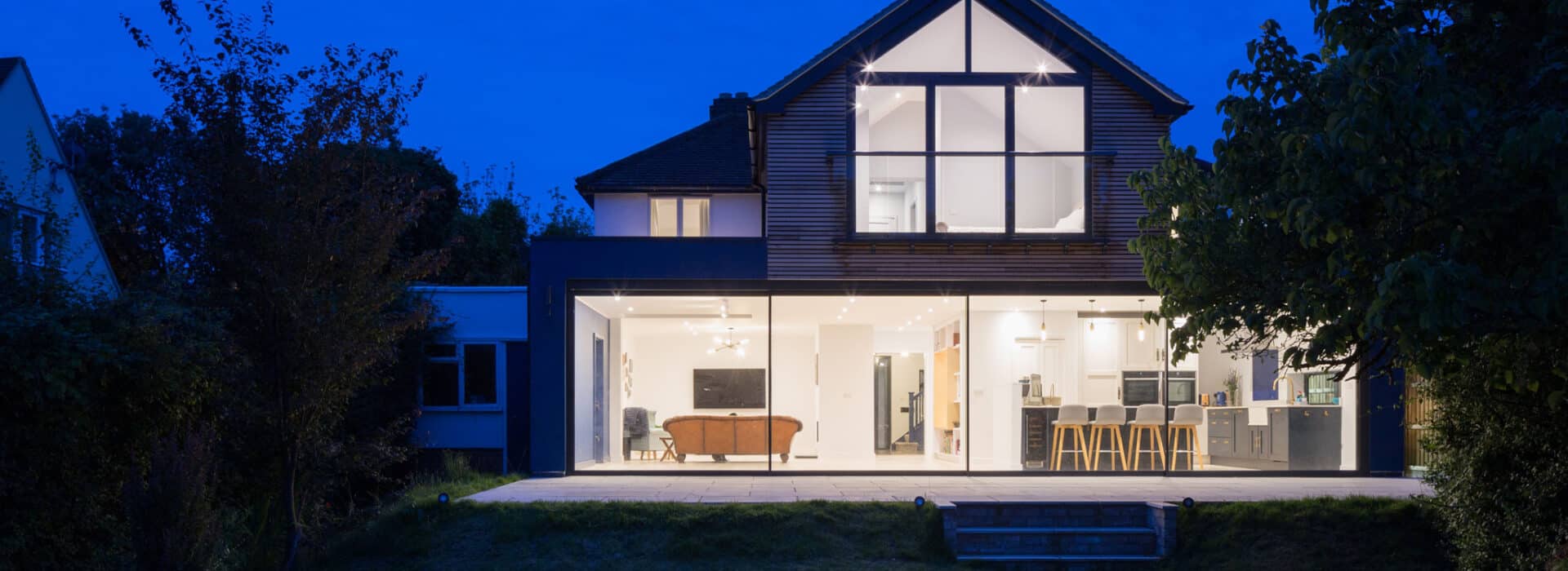 Modern two-story house with large windows and sliding doors, illuminated at night, featuring an open plan living area with furniture visible inside.